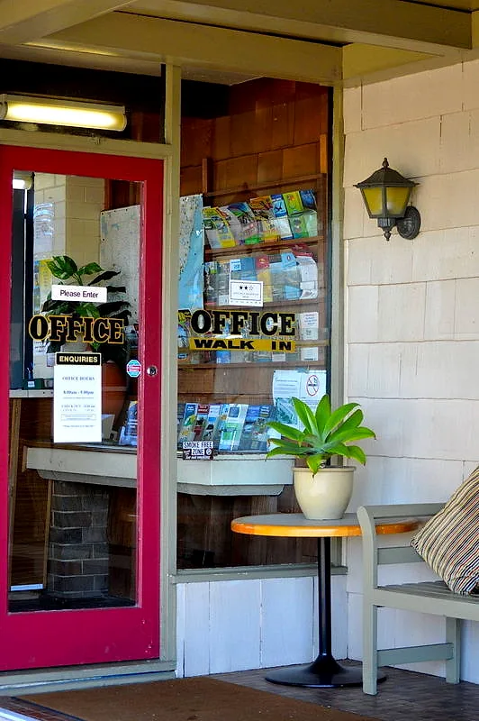 Mayfield Motel’s reception entrance — a red-framed glass door with gold OFFICE lettering, a brochure rack, timber panelling and a green bench seat.