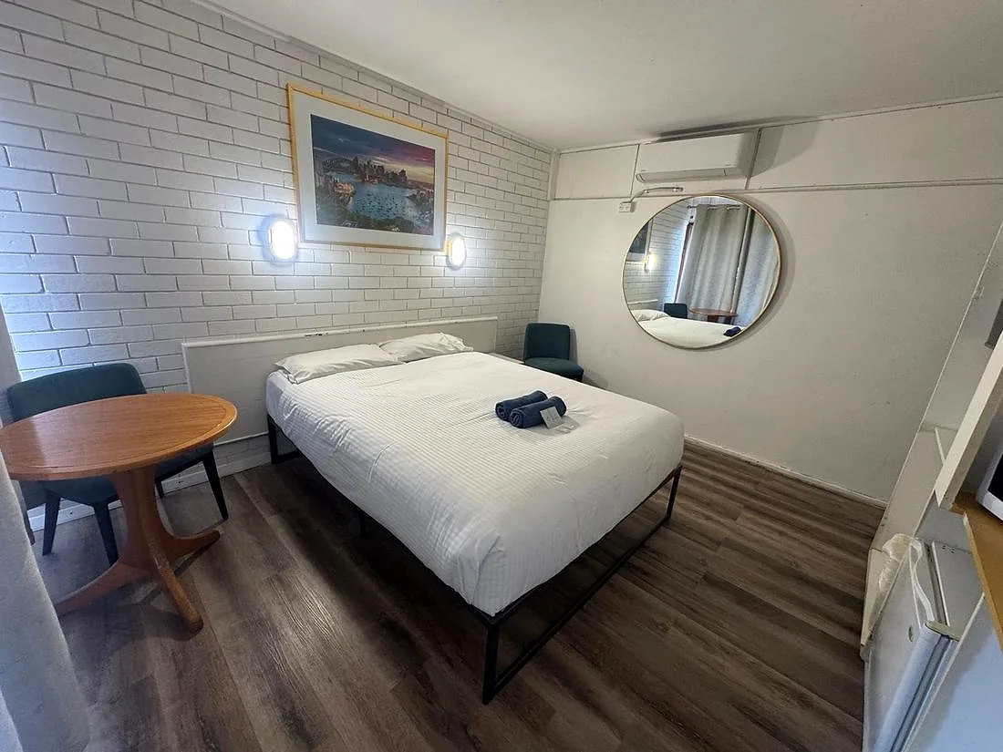 A refurbished room at Mayfield Motel — painted white brick, new timber-look flooring, a queen bed with fresh white linen, a round mirror and wall sconces.